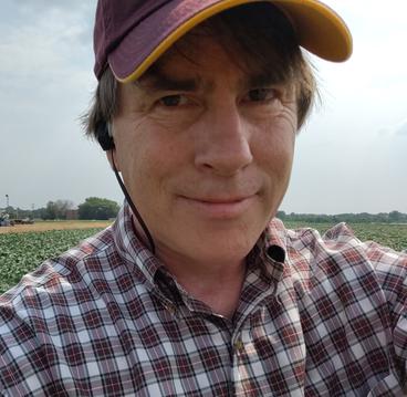 Photo of Jonathan Dregni. White male wearing button up shirt and a maroon and gold baseball cap, sanding in a soybean field.